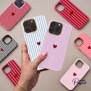 May include: A collection of smartphone cases featuring vertical stripes in various colours, including red, blue, pink, black, and purple. Each case displays a small, centred red heart icon. The cases have cutouts for camera lenses.
