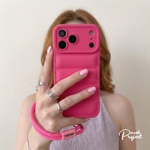May include: A bright pink puffer-style phone case with a quilted texture and a matching pink wrist strap featuring a small rectangular tag with the word TREND printed in black. The case has a raised camera cutout for three lenses and a flash.
