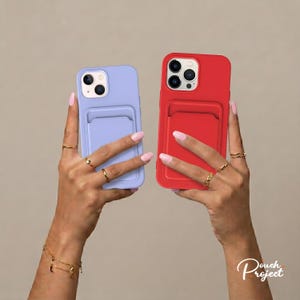 Peut inclure: Deux coques de téléphone, une lavande et une rouge, chacune avec un porte-cartes au dos. Les coques sont tenues par des mains avec des bagues et des bracelets. Les coques sont pour un smartphone.
