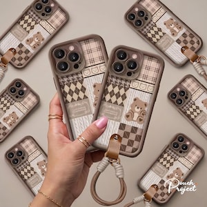 May include: Multiple phone cases featuring a patchwork design of brown and beige plaid, argyle, and cable knit patterns with cartoon bear illustrations. Each case includes a brown leather-like tab attached to a braided cord wrist strap.