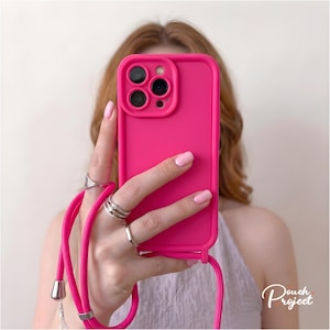 Könnte beinhalten: Eine leuchtend pinke Smartphone-Hülle mit passendem integriertem Umhängeband. Die Hülle verfügt über einen erhöhten Kameraausschnitt für ein Triple-Objektiv-System und eine glatte, matte Oberfläche. Eine silberfarbene Metallspitze ist am Band sichtbar.