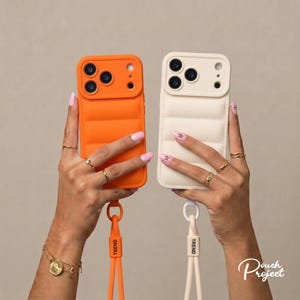 May include: Two puffer-style phone cases, one bright orange and one off-white, each featuring a raised camera cutout and a matching detachable wrist strap with the word TREND printed in black. The cases have a quilted, padded texture.
