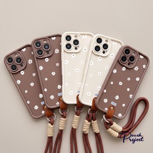 May include: Five smartphone cases arranged in a fan shape, featuring brown and cream backgrounds with a white daisy pattern. Each case includes a brown leather attachment point connected to a brown rope wrist strap with beige cord accents.