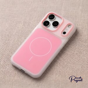 Pu&ograve; includere: Una custodia per telefono rosa e bianca con design MagSafe. La custodia ha un retro rosa con un cerchio bianco e una piccola linea e un bordo bianco. L'area della fotocamera ha tre lenti nere e un bordo rosa.