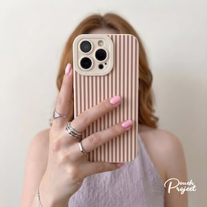 Puede incluir: Una funda para teléfono móvil con un patrón de rayas verticales en marrón claro y blanco. La funda incluye un protector de lente de cámara elevado en un tono marrón claro a juego con el texto Pro Camera impreso en el marco del módulo.