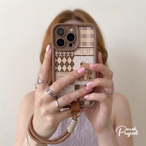 May include: A brown smartphone case featuring a patchwork design of plaid, argyle, and houndstooth patterns in shades of brown and cream. The case includes a cartoon bear illustration and a matching brown wrist strap with a beige cord attachment.