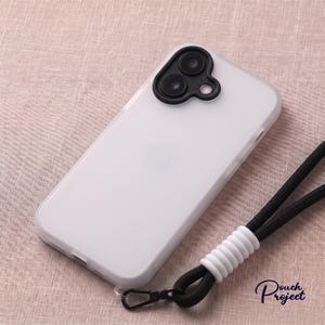 May include: A translucent white smartphone case featuring a black camera lens protector with two circular cutouts. A black woven lanyard is attached to the bottom corner, secured by a white ribbed plastic connector and a small black metal clip.