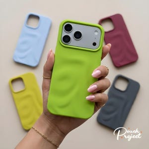 Puede incluir: Una mano sosteniendo una funda de teléfono verde lima con una textura ondulada, rodeada de otras fundas de teléfono en azul claro, amarillo, burdeos y gris. Las fundas tienen un acabado liso y mate y están diseñadas para un smartphone.