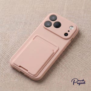 Può includere: Custodia per telefono rosa pallido con portacarte integrato. La custodia ha una finitura liscia e presenta tre obiettivi della fotocamera e un flash. Il logo Pouch Project è visibile in basso a destra.