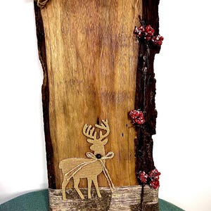May include: A rustic wooden wall hanging with a deer silhouette and red berry accents. The rectangular plaque features a natural wood grain, a bark edge, and a jute rope for hanging. The deer is tied with a jute bow.