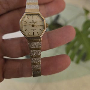 K&ouml;nnte beinhalten: Eine Vintage-Quarzuhr mit einem gold- und silberfarbenen Armband. Das Zifferblatt der Uhr ist achteckig mit goldenen Zeigern und Markierungen. Das Armband ist etwa 1,3 cm breit.