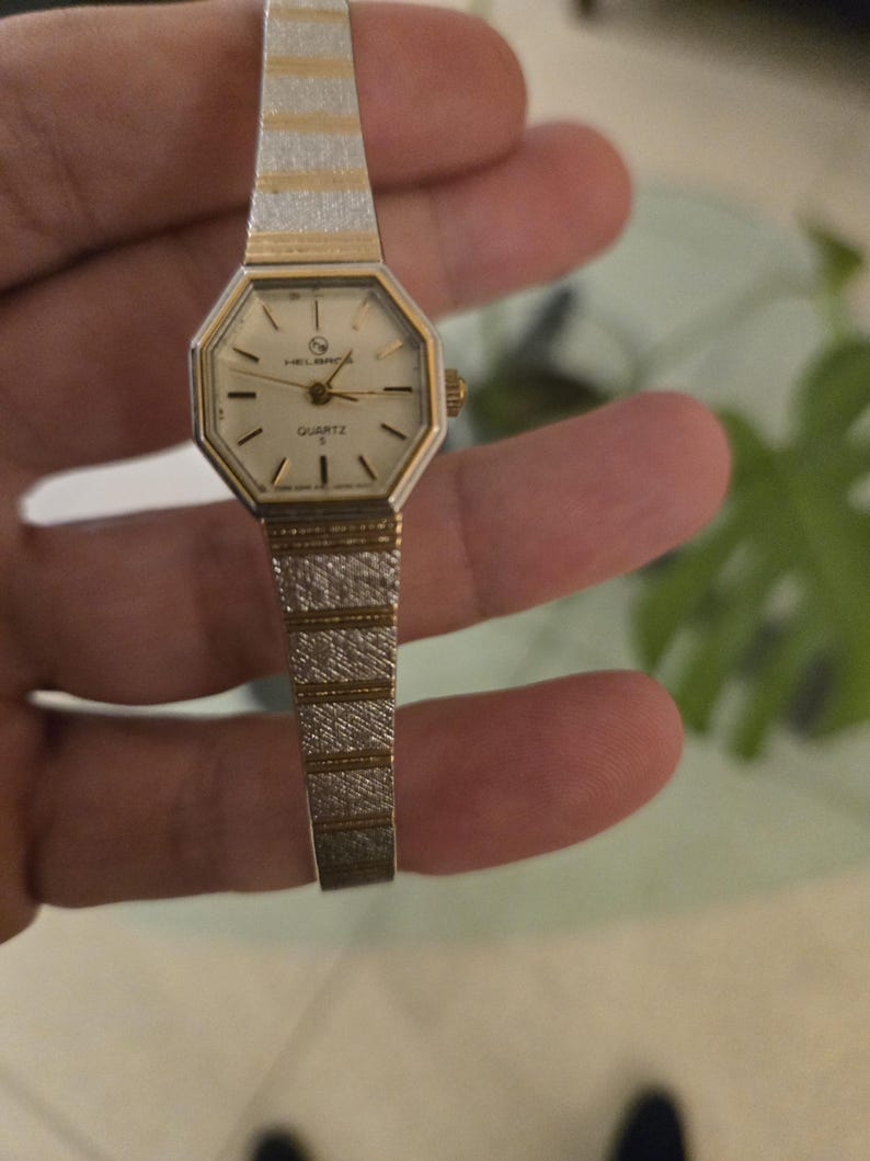 K&ouml;nnte beinhalten: Eine Vintage-Quarzuhr mit einem gold- und silberfarbenen Armband. Das Zifferblatt der Uhr ist achteckig mit goldenen Zeigern und Markierungen. Das Armband ist etwa 1,3 cm breit.