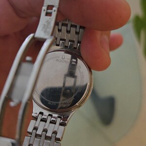 May include: A Bulova stainless steel wristwatch with a silver band and clasp. The watch face is not visible, but the back of the watch is visible, with the Bulova logo and text. The band is made of small, rectangular links.
