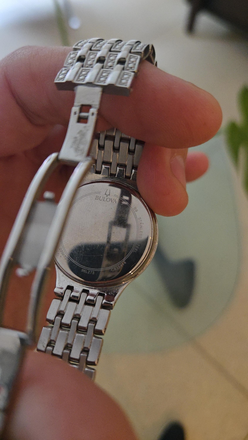 May include: A Bulova stainless steel wristwatch with a silver band and clasp. The watch face is not visible, but the back of the watch is visible, with the Bulova logo and text. The band is made of small, rectangular links.