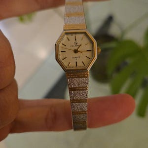 K&ouml;nnte beinhalten: Eine Vintage Helbros Quarzuhr mit einem goldfarbenen achteckigen Zifferblatt und einem silber- und goldfarbenen Armband. Das Zifferblatt hat schwarze Stundenmarkierungen und Zeiger. Die Uhr wird zwischen den Fingern gehalten.