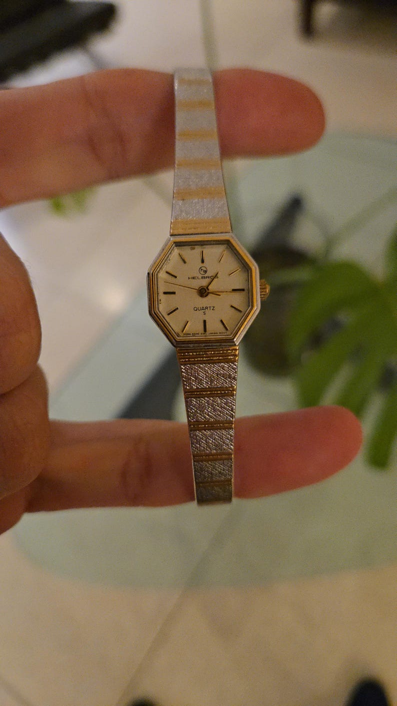 K&ouml;nnte beinhalten: Eine Vintage Helbros Quarzuhr mit einem goldfarbenen achteckigen Zifferblatt und einem silber- und goldfarbenen Armband. Das Zifferblatt hat schwarze Stundenmarkierungen und Zeiger. Die Uhr wird zwischen den Fingern gehalten.