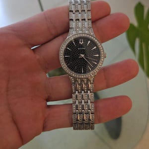 May include: A silver-toned Bulova watch with a black dial and a bezel and band adorned with small, sparkling crystals. The watch has a rectangular link bracelet and a round face. The watch is held in a hand.