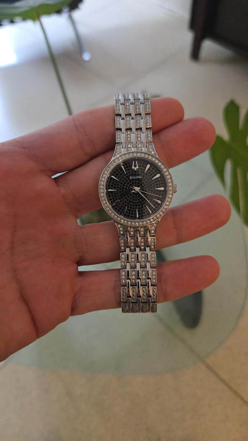 May include: A silver-toned Bulova watch with a black dial and a bezel and band adorned with small, sparkling crystals. The watch has a rectangular link bracelet and a round face. The watch is held in a hand.