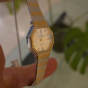 K&ouml;nnte beinhalten: Eine Vintage Helbros Quartz-Uhr mit silbernem und goldfarbenem Armband und cremefarbenem Zifferblatt. Die Uhr hat eine geometrische achteckige Form und ein zartes, strukturiertes Armband. Das Zifferblatt zeigt das Wort "QUARTZ".