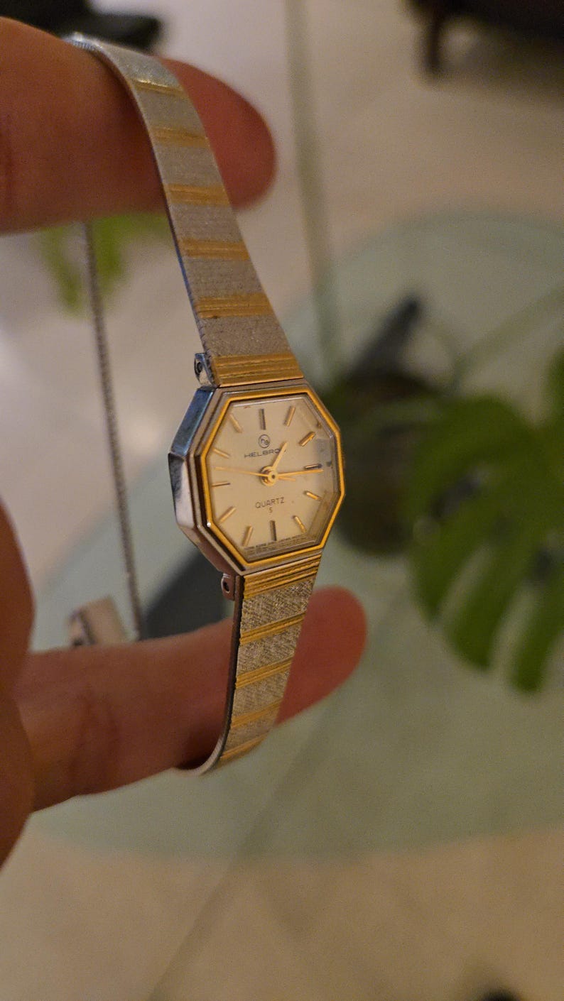 K&ouml;nnte beinhalten: Eine Vintage Helbros Quartz-Uhr mit silbernem und goldfarbenem Armband und cremefarbenem Zifferblatt. Die Uhr hat eine geometrische achteckige Form und ein zartes, strukturiertes Armband. Das Zifferblatt zeigt das Wort "QUARTZ".