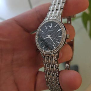 May include: A Bulova wristwatch with a silver-toned band and a dark face. The watch face is surrounded by a ring of small, sparkling stones. The band is made of interconnected rectangular links, also adorned with stones.