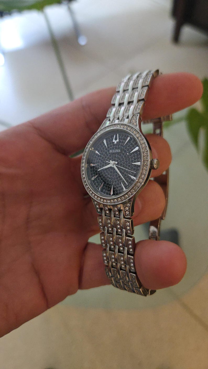 May include: A Bulova wristwatch with a silver-toned band and a dark face. The watch face is surrounded by a ring of small, sparkling stones. The band is made of interconnected rectangular links, also adorned with stones.