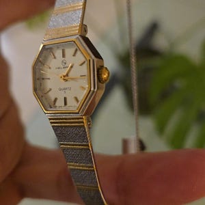 K&ouml;nnte beinhalten: Eine Vintage Helbros Quarzuhr mit einem goldfarbenen achteckigen Geh&auml;use und einem silber- und goldfarbenen Armband. Das Zifferblatt der Uhr ist wei&szlig; mit goldenen Zeigern und Markierungen. Das Armband hat ein strukturiertes Design.