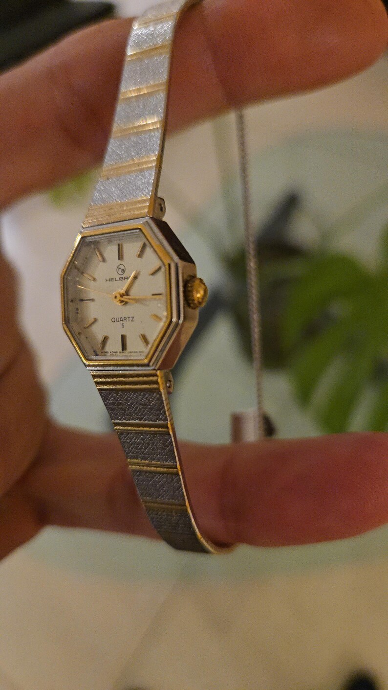 K&ouml;nnte beinhalten: Eine Vintage Helbros Quarzuhr mit einem goldfarbenen achteckigen Geh&auml;use und einem silber- und goldfarbenen Armband. Das Zifferblatt der Uhr ist wei&szlig; mit goldenen Zeigern und Markierungen. Das Armband hat ein strukturiertes Design.