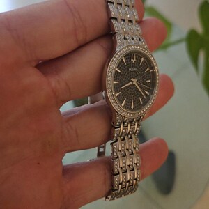 May include: A Bulova wristwatch with a silver-tone band and a dark face. The watch features a bezel and band adorned with small, sparkling crystals. The watch is held in a hand, showcasing its design and details.