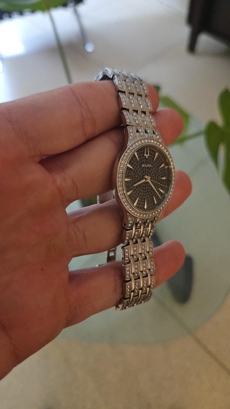 May include: A Bulova wristwatch with a silver-tone band and a dark face. The watch features a bezel and band adorned with small, sparkling crystals. The watch is held in a hand, showcasing its design and details.