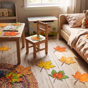 May include: A children's bedroom scene featuring a wooden table, chair, and bed decorated with numerous colourful paper maple leaves in shades of yellow, orange, red, and green. A teddy bear sits on the bed holding a leaf, with books and blocks nearby.