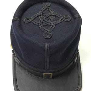 Puede incluir: Un kepi azul marino con visera de cuero negro y un diseño decorativo de cordón negro en la parte superior. El sombrero presenta una pequeña hebilla de latón y es probablemente un accesorio histórico o de estilo militar.