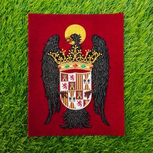 May include: Embroidered heraldic crest on a red background. The crest features a golden crown, a black eagle with a golden halo, and a shield with various emblems. The wings of the eagle are black. The background is a deep red.