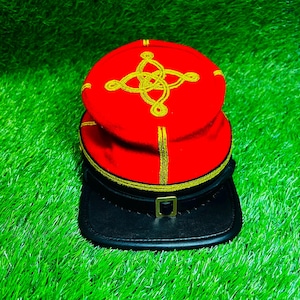 May include: A bright red military-style cap featuring a prominent gold embroidered knot design on the top crown. The cap includes gold piping trim, a black band with a central gold-toned buckle, and a black visor extending from the front base.