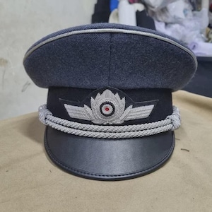 May include: A dark blue military-style hat with a rounded crown and a black visor. The hat features a silver emblem with a red center and silver rope detailing around the base of the crown.
