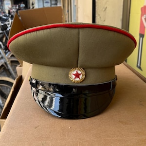 May include: An olive green military hat with a red trim and a glossy black peak. It has a gold-coloured badge with a red star. The hat is displayed on a cardboard surface.