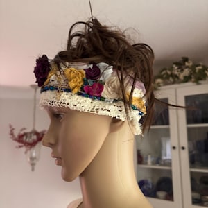 May include: A decorative headband featuring a layered arrangement of fabric flowers in shades of deep purple, mustard yellow, and cream. The band incorporates white lace trim, thin strands of blue and green beads, and small metallic accents.