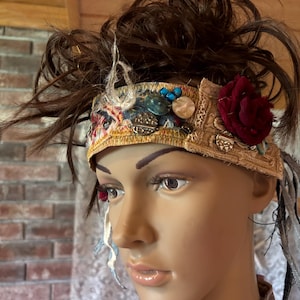 May include: A decorative fabric headband featuring a layered design with a dark red fabric flower, various metallic and blue circular embellishments, and frayed edges. The band is constructed from multiple textures of tan, blue, and patterned fabric.