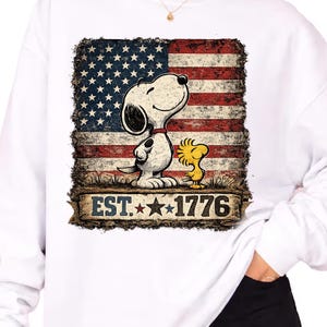 May include: A white long-sleeve sweatshirt featuring a graphic print of Snoopy and Woodstock standing in front of a distressed American flag. Below the characters, a banner displays the text EST. 1776 with two stars between the numbers.