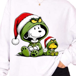 Peut inclure: Un sweat-shirt blanc à manches longues avec une impression graphique représentant un chien blanc et un oiseau jaune portant des tenues vertes à capuche et des bonnets de Noël rouges. Ils ont des expressions grincheuses et des pyjamas verts.