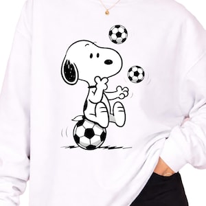 May include: White sweatshirt featuring a black and white cartoon illustration of Snoopy balancing on a soccer ball while juggling two more. The design is centered on the front of the long-sleeved top. The sweatshirt has a crew neck.