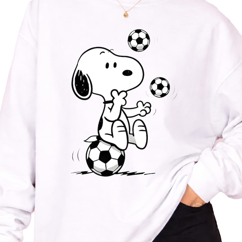 May include: White sweatshirt featuring a black and white cartoon illustration of Snoopy balancing on a soccer ball while juggling two more. The design is centered on the front of the long-sleeved top. The sweatshirt has a crew neck.