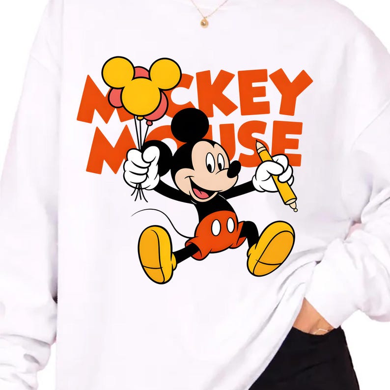 May include: A white long-sleeve sweatshirt featuring a graphic of Mickey Mouse holding a yellow pencil and a cluster of yellow balloons. The text MICKEY MOUSE is printed in large orange letters behind the character. The design is centered on the chest.