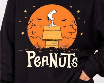 Puede incluir: Una sudadera negra de manga larga con un gráfico de Snoopy sentado en su caseta frente a una gran luna naranja. El diseño incluye siluetas de árboles desnudos, murciélagos y estrellas. La palabra PEANUTS aparece en grandes letras de color crema.
