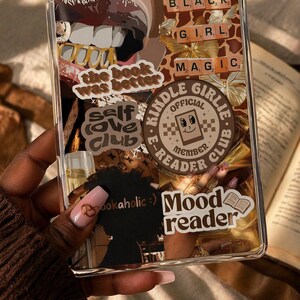 May include: A clear e-reader case decorated with various stickers including text that reads the book was better, self love club, Kindle Girlie E-Reader Club Official Member, bookaholic, and Mood reader, alongside images of lips, butterflies, and gold.