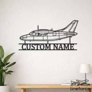 May include: A black metal wall art piece featuring a side-profile silhouette of a high-wing airplane. Below the aircraft, a horizontal bar contains the text CUSTOM NAME in a bold, sans-serif font, supported by two vertical metal rods.