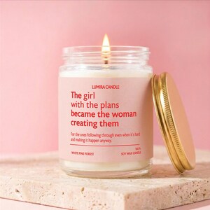 May include: A lit soy wax candle in a glass jar with a gold lid. The candle has a pink label with the text "The girl with the plans became the woman creating them." The candle is labeled "White Pine Forest."