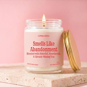 May include: A lit soy wax candle in a glass jar with a gold lid. The label reads "Smells Like Abandonment" with additional text. The candle is on a stone surface against a pink background. The candle is labeled "White Pine Forest".