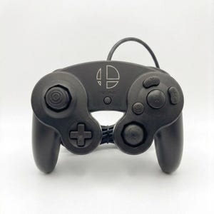 May include: A black wired video game controller featuring the Super Smash Bros. logo in the centre. The controller includes a large central A button, smaller B, X, and Y buttons, a directional pad, two analogue sticks, and a Start/Pause button.