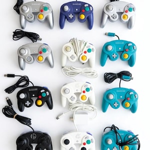 May include: A collection of twelve wired video game controllers arranged in a grid. The controllers feature various colours including silver, indigo, black, white, and teal, each with a central analogue stick, D-pad, and coloured action buttons.